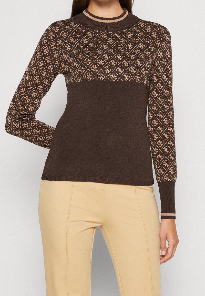 Femme portant un pull brun à manches longues avec un motif géométrique beige sur la poitrine supérieure et les manches, associé à un pantalon beige.