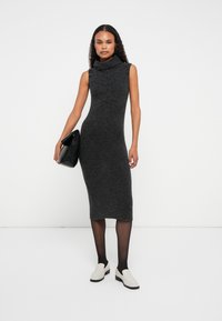 Αμάνικο, σκούρο γκρι πλεκτό φόρεμα με ψηλό λαιμό, υφή μπροστά, συνδυασμένο με μαύρες κολάν και ανοιχτόχρωμα παντόφλες.