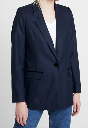 Kobieta ubrana w ciemnogranatową marynarkę na jeden guzik, pod którą ma biały top i niebieskie dżinsy, stoi na tle jednolitego, jasnoszarego tła.