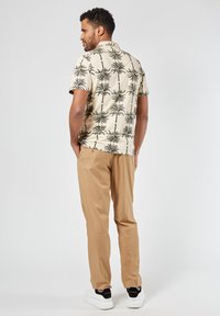Korte mouwen, crèmekleurig shirt met zwart palmbomenpatroon, gecombineerd met lichtbeige broeken. De persoon draagt witte sneakers met zwarte accenten.