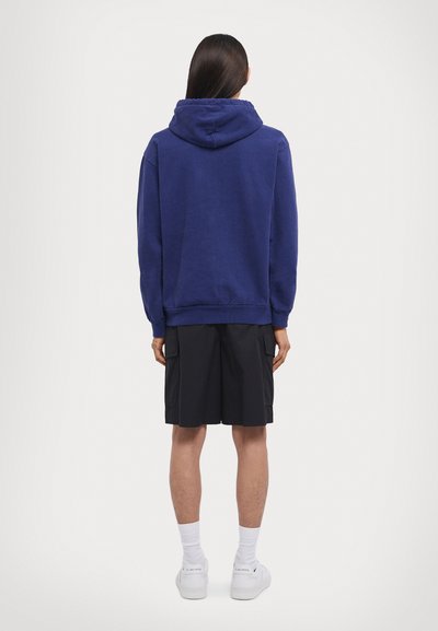 Lila Kapuzenpullover mit weichem Material und Schnürung an der Kapuze, kombiniert mit schwarzen Cargo-Shorts und weißen Sneakers, von hinten betrachtet.