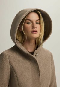 Brązowy płaszcz z wełny z dużym kapturem, z wysoką kołnierzem i zapięciem na zatrzaski. Gładka tekstura i minimalistyczny design.