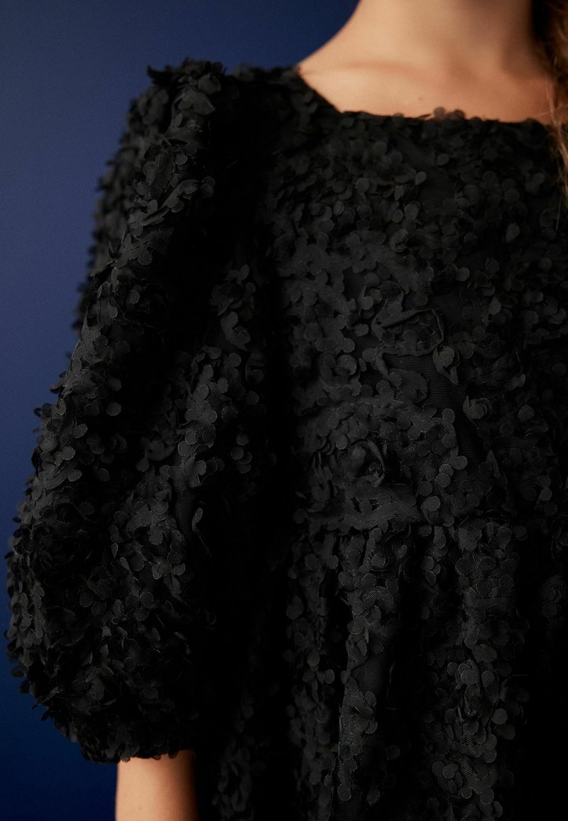 Robe noire en tissu texturé avec des applications florales en relief, des manches bouffantes et un décolleté carré, mettant en valeur des détails complexes.