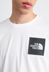 Weißes Baumwoll-T-Shirt mit einem schwarzen quadratischen Logo und dem Text "THE NORTH FACE" sowie einer geschwungenen Grafik. Kurze Ärmel und Rundhalsausschnitt.