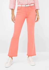 Koraalkleurige flare jeans hebben een hoge taille, rafelige zomen en worden gecombineerd met een witte riem en sneakers voor een casual uitstraling.