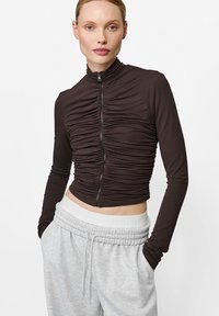 Brązowa, długozasuwana bluza z zebranym teksturą, skróconym krojem i wysokim kołnierzem; noszona z szarymi spodniami dresowymi na sznurek.