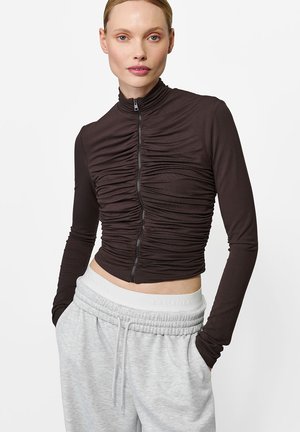 Brązowa, długozasuwana bluza z zebranym teksturą, skróconym krojem i wysokim kołnierzem; noszona z szarymi spodniami dresowymi na sznurek.