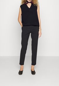 Svart ärmlös stickad tröja med V-hals, kombinerad med svarta skräddarsydda byxor. Skorna är svarta, platta, med en texturerad yta.