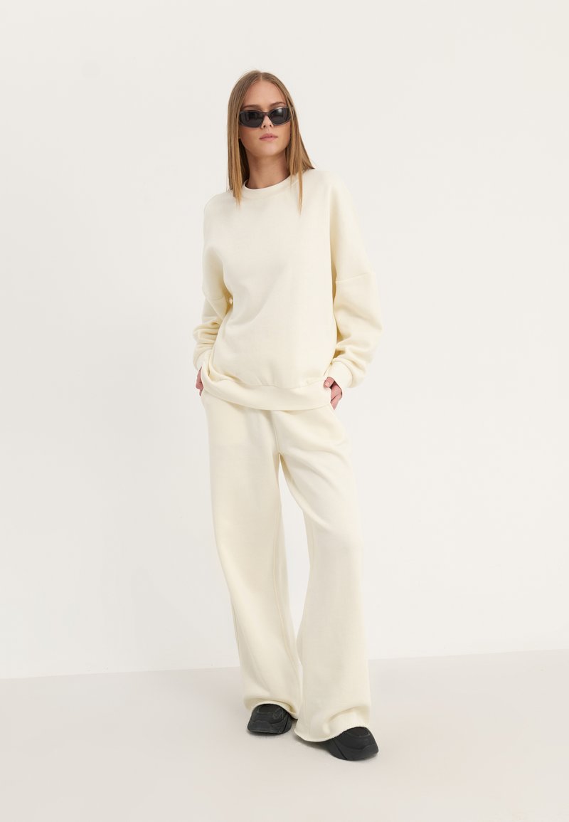 Femme portant un sweat-shirt et un pantalon oversize couleur crème, avec des chaussures noires et des lunettes de soleil, debout devant un fond clair uni.