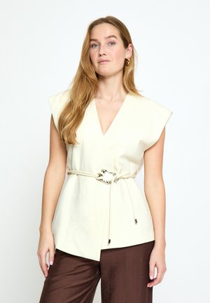 Pikajuustega blond naine, kes kannab varrukateta kremi värvi seotavat topi õhukese vööga ja pruune pükse, seisab tavalise valge tausta ees.