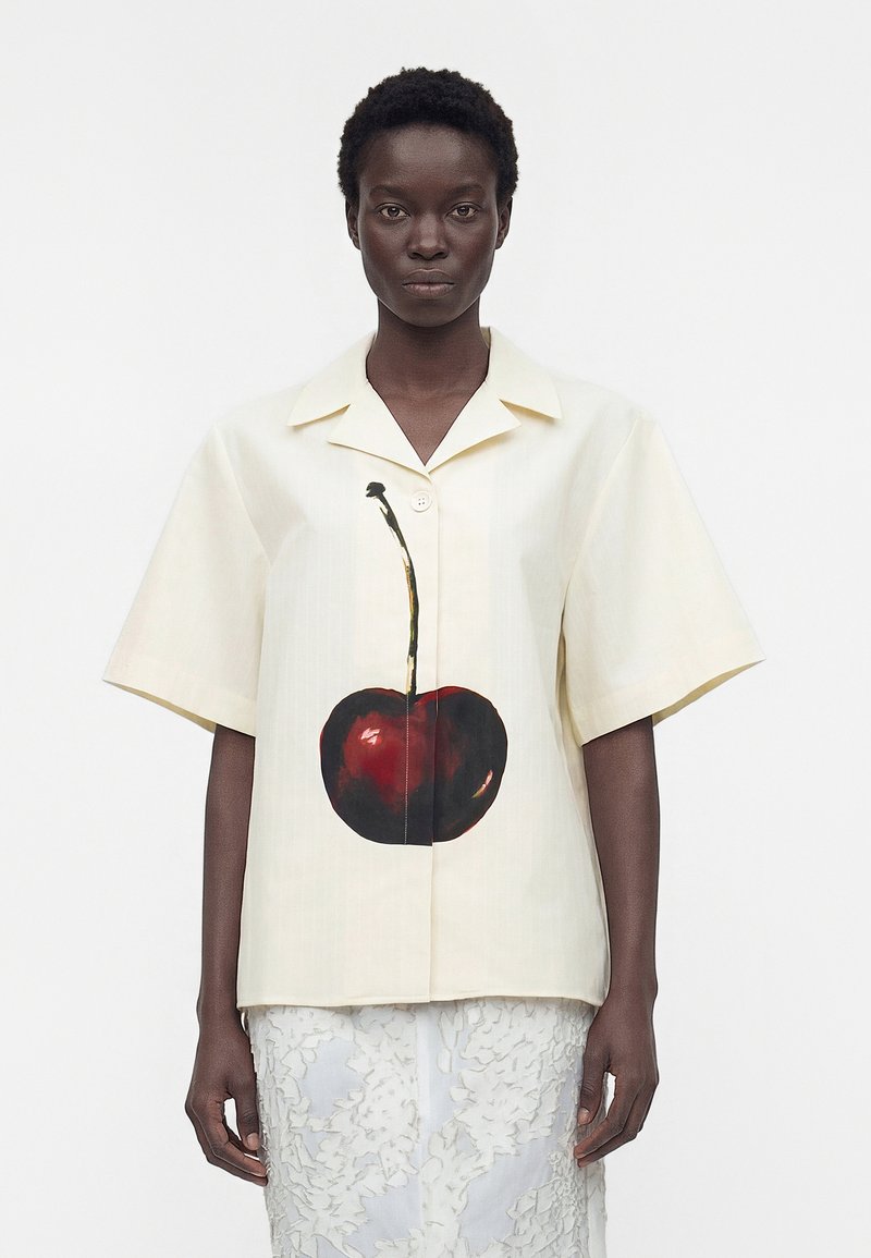 Γυναίκα φοράει μια κρεμ κοντομάνικη πουκάμισο με μεγάλα κόκκινα κεράσια στο μπροστινό μέρος και μια λευκή υφασμάτινη φούστα.