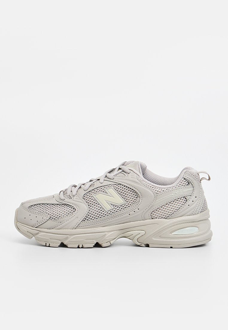 Zapatilla deportiva de color gris claro con materiales de malla y cuero, punta redonda, suela gruesa y un logotipo prominente en el lateral. Diseño de cordones.