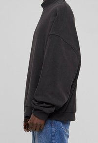 Sudadera oscura de gran tamaño con hombros caídos, usada por una persona de perfil, combinada con vaqueros azules sobre un fondo liso.