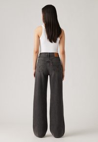 Jean taille haute à jambe large en denim gris foncé, avec des poches arrière ornées de coutures et un patch en simili cuir beige au niveau de la taille.
