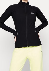 Svart idrottsjacka med hög krage, dragkedja framtill och två sidofickor. Har DKNY-logotypen på bröstet och en slimmad, figurnära design.