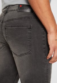 Jean en denim gris foncé montrant un gros plan de la poche arrière et de l'étiquette de la marque. Texture usée avec une coupe ajustée.