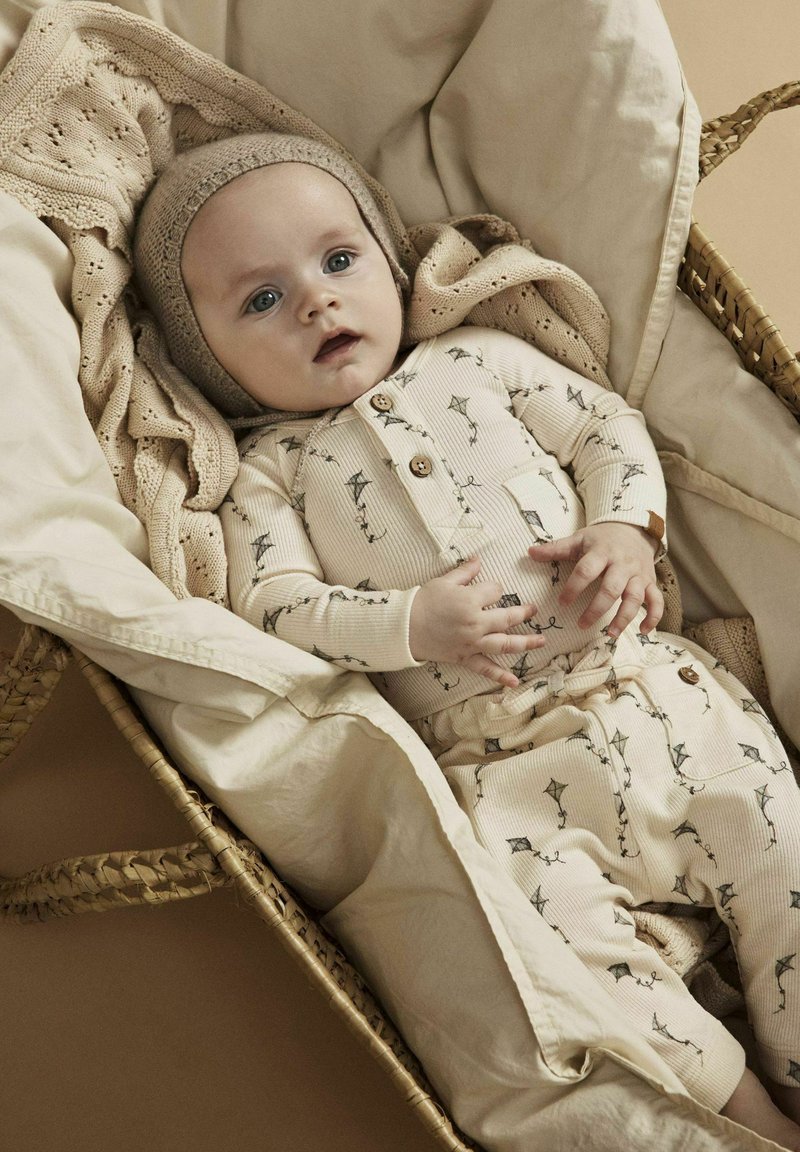 Helles beiges Babyoutfit mit Drachenmuster, bestehend aus einem knöpfbaren Oberteil und passenden Hosen. Eine gestrickte Mütze und eine strukturierte Decke sind ebenfalls enthalten.