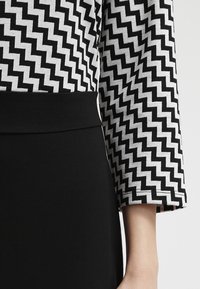 Robe noir et blanc avec un tissu à motif zigzag sur le haut et une jupe noire unie. Les manches sont de longueur trois-quarts avec une finition soignée.