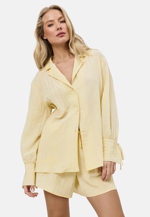 Mujer rubia vistiendo una camisa suelta de manga larga y color amarillo pálido con botones y pantalones cortos a juego, de pie frente a un fondo blanco.