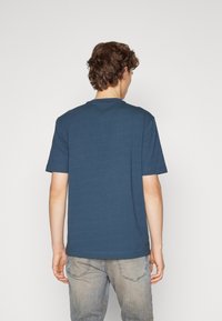 Krátký rukáv, tričko s kulatým výstřihem v tmavě modré tkanině, s hladkou texturou a rovnými lemy. Nosič má na sobě světlé džíny.