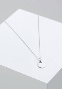 Collier en chaîne argentée avec un petit pendentif rond et lisse reposant sur une surface géométrique blanche.