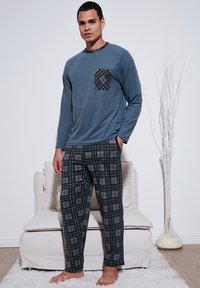 Kék hosszúujjú felső, mintás zsebbe, fekete kockás pizsama nadrággal párosítva. Simogató anyag, laza szabás, mezítláb viselve.