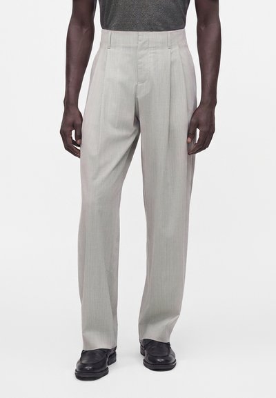 Homme portant un pantalon plissé gris clair avec des chaussures noires sur un fond blanc uni, montré de la moitié du torse jusqu’aux pieds.