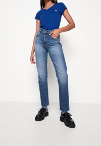 Kvinna som bär en blå kortärmad topp, högmidjade urtvättade blå jeans och svarta kraftiga snörskor, stående mot en vit bakgrund.