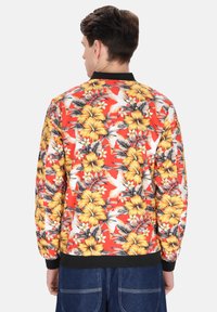 Bomber jacket floreale rossa con grandi fiori di ibisco gialli, foglie verdi e polsini neri. Realizzata in un tessuto liscio e leggero.