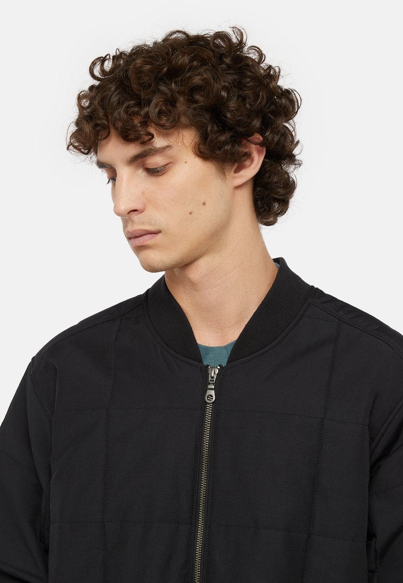 Veste bomber noire avec un design matelassé, col côtelé et fermeture éclair. Caractéristiques d'une texture lisse et d'une silhouette minimaliste.
