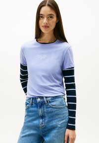T-shirt corto a maniche corte di colore lilla sovrapposto a una camicia a maniche lunghe a righe blu e bianche, abbinato a jeans in denim blu a vita alta.
