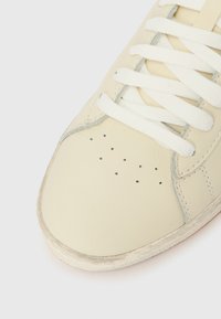 Αθλητικό παπούτσι εκρού χρώματος με λευκά κορδόνια, διάτρητη περιοχή στα δάχτυλα και ελαφρώς φθαρμένη σόλα πάνω σε λευκό φόντο.