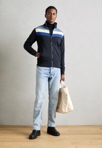 Jachetă navy cu fermoar, cu dungi orizontale albastre și crem, asociată cu blugi albaștri deschis și cizme chunky negre. Modelul ține o geantă tote bej.