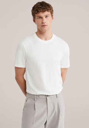 Jonge man met krullend haar, gekleed in een effen wit T-shirt met korte mouwen en lichtgrijze broek, staand tegen een neutrale achtergrond.