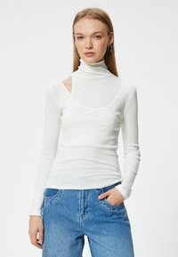 Weißes Langarmoberteil mit hohem Kragen und Ausschnitt an den Schultern, aus einem dünnen, glatten Stoff, kombiniert mit blauen Jeans.