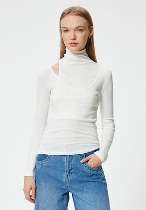 Weißes Langarmoberteil mit hohem Kragen und Ausschnitt an den Schultern, aus einem dünnen, glatten Stoff, kombiniert mit blauen Jeans.