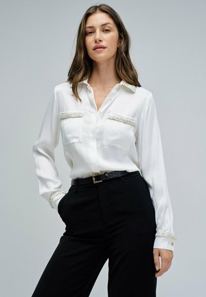 Mujer vistiendo una blusa blanca de manga larga con un borde texturizado en los bolsillos y los puños, combinada con pantalones negros y un cinturón negro, de pie frente a un fondo gris.