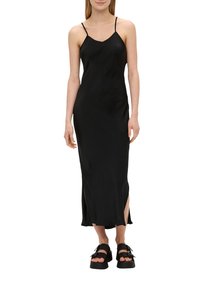 Schwarzes, ärmelloses Midikleid mit dünnen Trägern, das seitliche Schlitze und eine glatte Textur aufweist. Kombiniert mit klobigen schwarzen Plateauschuhen.