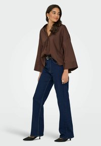 Femme portant une chemise ample marron à boutons, un jean bleu foncé, une ceinture noire, des talons pointus noirs, debout devant un fond clair uni.