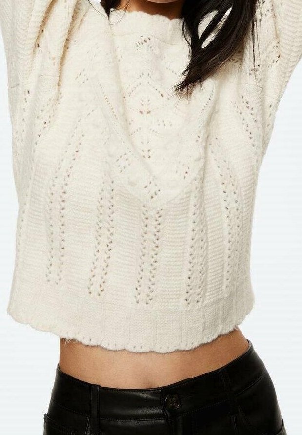 Pull court blanc avec motif en maille texturée, présentant des détails en points ouverts et un ourlet festonné, associé à un pantalon noir taille haute.