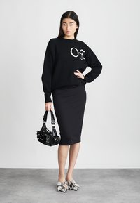 Vrouw in een zwarte sweater met de tekst "Off", zwarte kokerrok, houdt een zwart gestikte handtas vast en draagt zilveren puntige schoenen met strikken.