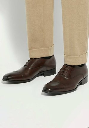 Bruine leren nette schoenen met een puntige neus, voorzien van vijf veterogen en een strak ontwerp. Gecombineerd met beige opgerolde pantalons.