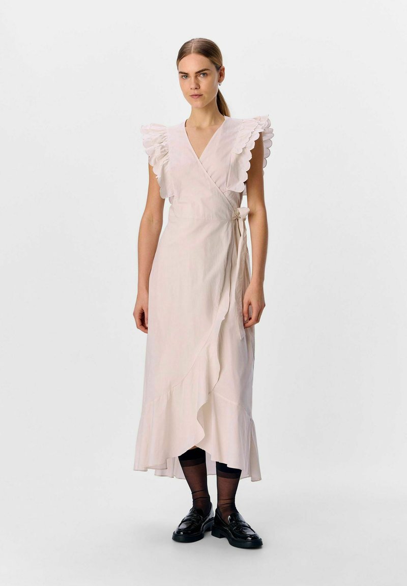 Robe portefeuille rose clair avec des manches en dentelle, un ourlet volé et une taille à nouer. Portée avec des chaussures noires et des collants noirs transparents. Texture lisse.