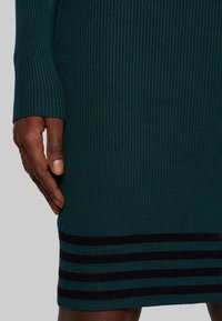 Abito in maglia di un verde scuro con una texture a coste. Presenta un orlo con strisce orizzontali nere. Dettaglio ravvicinato della manica e della posizione della mano.