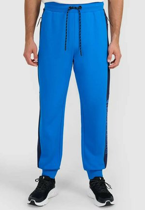 Mężczyzna noszący jasnoniebieskie spodnie dresowe z czarnymi bocznymi panelami oraz czarne buty sportowe z białymi podeszwami.