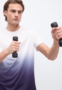 Νεαρός άνδρας με λευκό και μοβ πουδρένιο πουκάμισο κρατώντας και σηκώνοντας δύο μικρούς μαύρους αλτήρες μπροστά από ένα απλό λευκό φόντο.