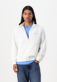 Lightweight white jacket with a high collar, elastic hem, and Tommy Jeans logo. Worn over a blue shirt and paired with blue jeans.