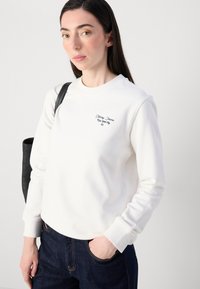 En kvinne med langt mørkt hår poserer, iført en hvit sweatshirt med logodetaljer, sammen med mørke jeans og en svart skulderveske.