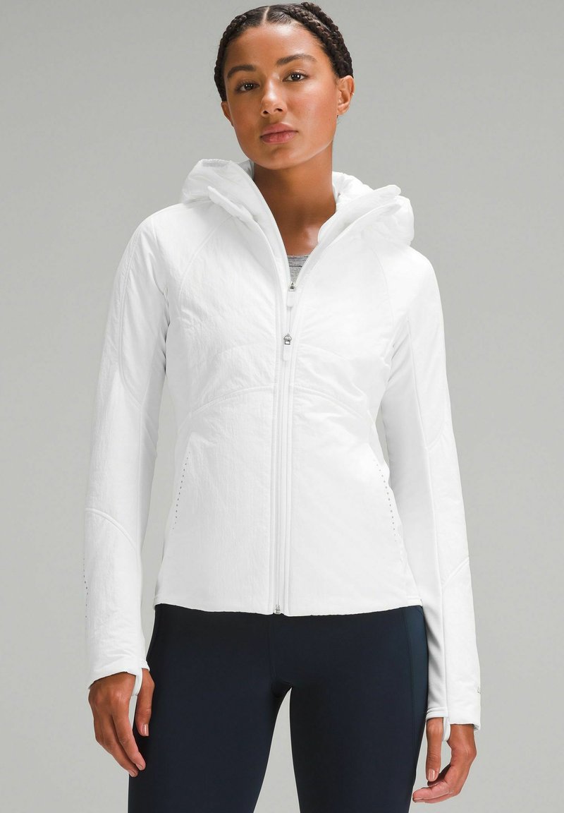 Veste légère blanche à fermeture éclair avec capuche, dotée d'une finition texturée et de passes-pouces dans les manches. Associée à des leggings navy.