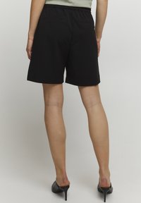 Pantaloni corti neri con cintura elastica, due tasche posteriori e una superficie in tessuto liscia. Il modello indossa tacchi neri.